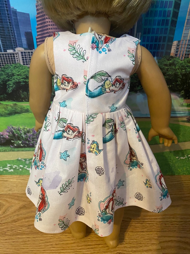 May include: A pink sleeveless dress with a mermaid print. The dress features Ariel, Flounder, and Sebastian from the Disney film The Little Mermaid.