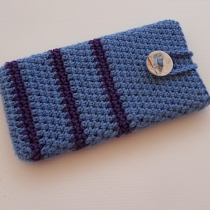 Könnte beinhalten: Eine blaue gehäkelte Handyhülle mit lila Streifen und einem Knopfverschluss. Der Knopf zeigt ein Vogelmotiv.