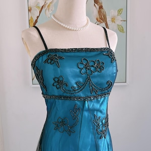 May include: A teal formal dress with black beaded floral embroidery. The dress features spaghetti straps, a square neckline, and a flowing skirt. A pearl necklace is worn around the mannequin's neck.