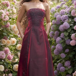 May include: A strapless, floor-length, burgundy gown with a fitted bodice and a flowing skirt. The bodice is adorned with delicate embroidery and the skirt has a subtle sheen.