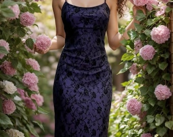 May include: A woman wearing a long, dark purple dress with black lace detailing. The dress is sleeveless and has a fitted silhouette. The woman is standing in front of a wall of pink flowers.