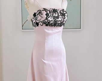 May include: A light pink formal gown with a fitted bodice and a flowing skirt. The bodice features a white panel with intricate black floral embroidery. The dress has thin spaghetti straps and a pearl necklace. The gown is displayed on a mannequin.