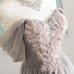 May include: A gray tulle gown with a sweetheart neckline and off-the-shoulder sleeves. The bodice is adorned with delicate floral embroidery in a blush pink color.