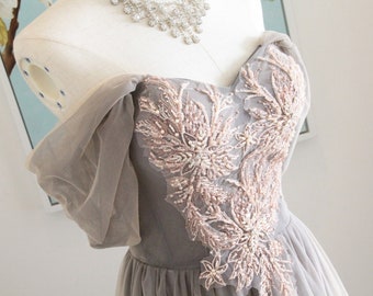 May include: A gray tulle gown with a sweetheart neckline and off-the-shoulder sleeves. The bodice is adorned with delicate floral embroidery in a blush pink color.