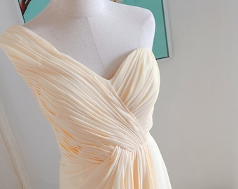 May include: A cream-colored, one-shoulder formal dress with a sweetheart neckline. The bodice features intricate pleating that flows into a draped design. The dress is displayed on a mannequin, showcasing its elegant design.