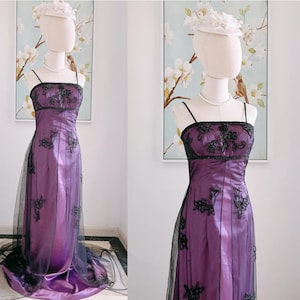 May include: A formal purple gown with black floral embroidery and spaghetti straps. The dress features a sheer black overlay and is displayed on a mannequin. A white hat and pearl necklace complete the look. The dress is suitable for a special occasion.