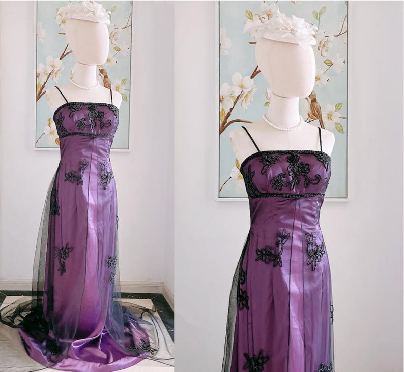 May include: A formal purple gown with black floral embroidery and spaghetti straps. The dress features a sheer black overlay and is displayed on a mannequin. A white hat and pearl necklace complete the look. The dress is suitable for a special occasion.