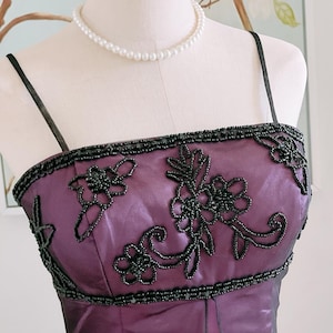 May include: A formal purple dress with black beaded floral embellishments. The dress features thin black straps and a beaded neckline. A pearl necklace is draped around the mannequin's neck.
