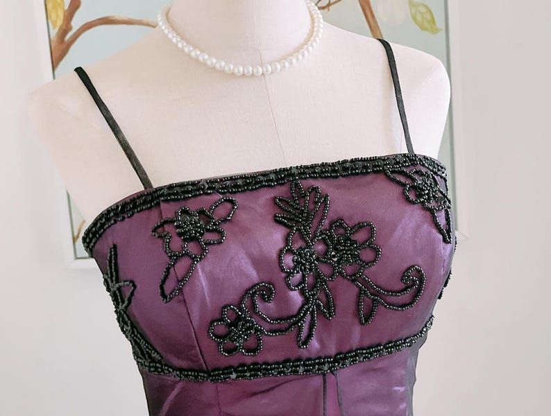 May include: A formal purple dress with black beaded floral embellishments. The dress features thin black straps and a beaded neckline. A pearl necklace is draped around the mannequin's neck.