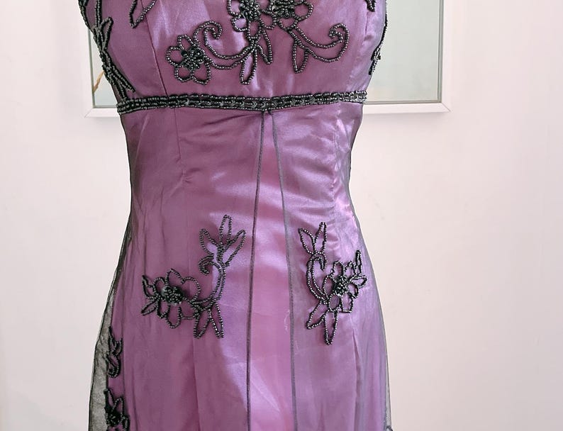 May include: A formal, strapless, lavender-colored dress. The bodice and waistline are embellished with dark gray beading. Floral beaded designs cascade down the front of the dress. The dress is made of a shiny, silky material and has a sheer overlay.