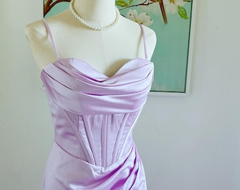 Pode incluir: Um vestido de cetim lavanda com decote coração e um corpete estilo corset. O vestido tem uma silhueta ajustada e uma saia fluida.