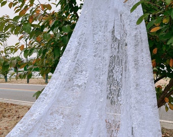 May include: White lace bridal cape with a hood and a bow tie. The cape is hanging on a tree branch.