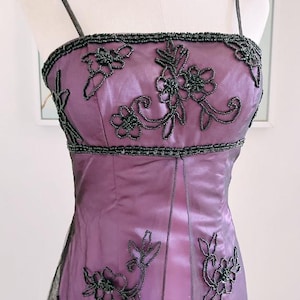 May include: A formal, floor-length purple dress with black beaded floral embellishments. The dress features a square neckline, spaghetti straps, and a fitted bodice. A pearl necklace is also visible.