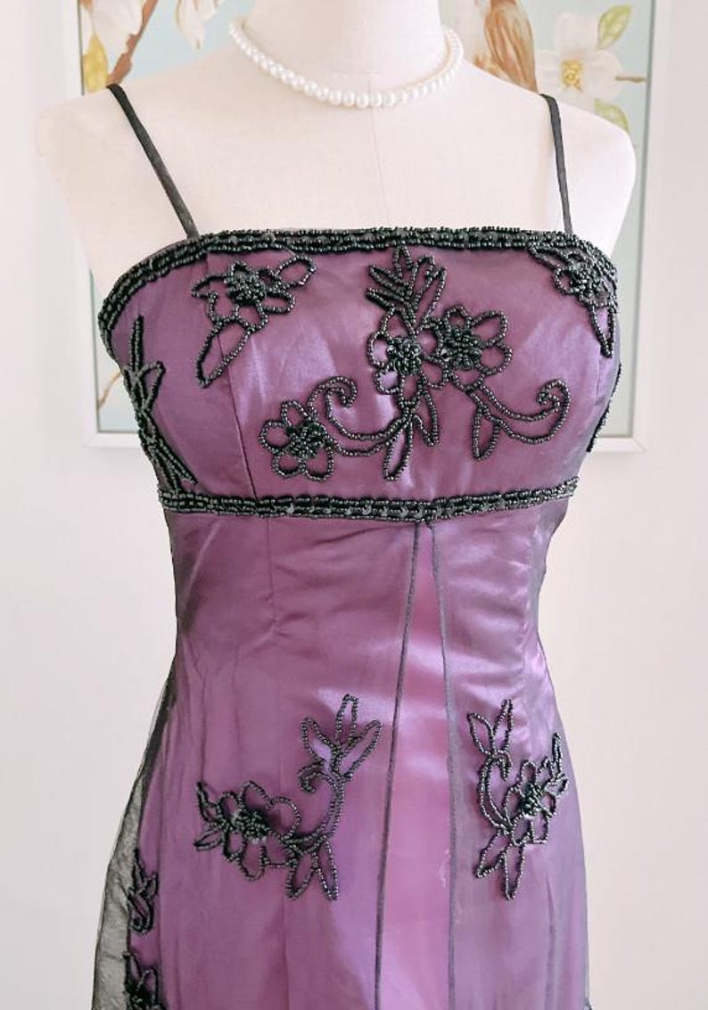 May include: A formal, floor-length purple dress with black beaded floral embellishments. The dress features a square neckline, spaghetti straps, and a fitted bodice. A pearl necklace is also visible.