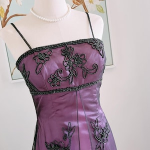 May include: A formal purple dress with black beaded floral embellishments. The dress features a square neckline and thin straps. The bodice and hemline are adorned with intricate floral beadwork. A pearl necklace is also visible.