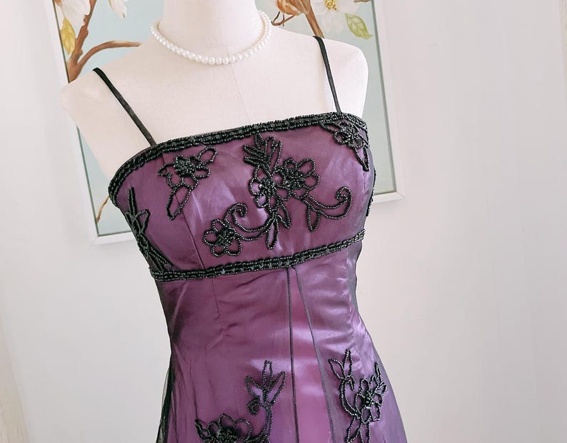 May include: A formal purple dress with black beaded floral embellishments. The dress features a square neckline and thin straps. The bodice and hemline are adorned with intricate floral beadwork. A pearl necklace is also visible.