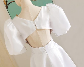 Pode incluir: Um vestido branco de cetim com costas baixas e mangas bufantes. O vestido tem um corpete justo e uma saia fluida.