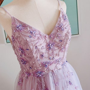 May include: A lavender-colored tulle dress with a V-neckline and delicate floral embroidery. The dress is adorned with purple and silver beads and sequins.