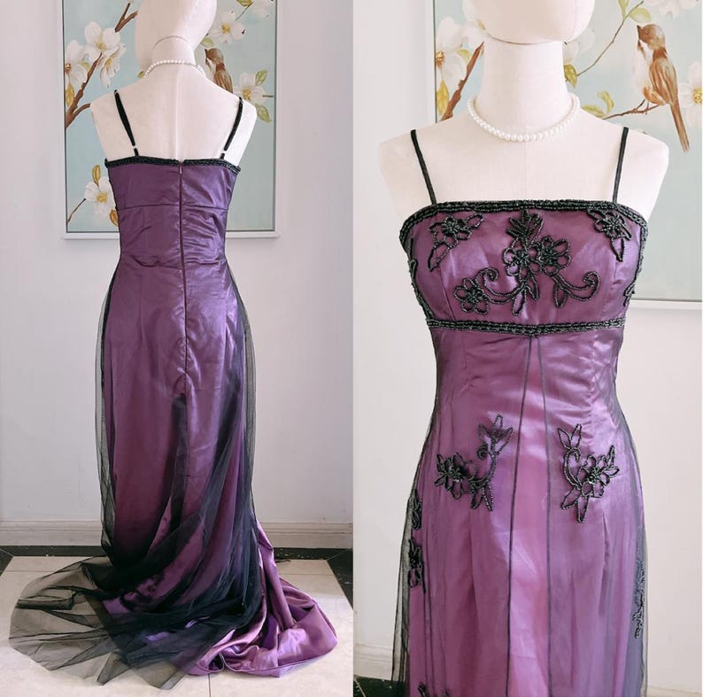 May include: A formal purple gown with black beaded accents and spaghetti straps. The bodice features intricate floral beadwork, and the skirt is overlaid with a sheer black fabric that trails behind the dress. The dress is displayed on a mannequin.