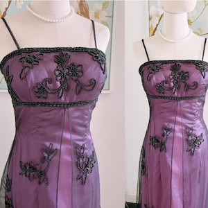 May include: A formal purple dress with black beaded floral embellishments. The dress features a square neckline, spaghetti straps, and a fitted bodice. The skirt is a flowing style, and the dress is displayed on a mannequin.