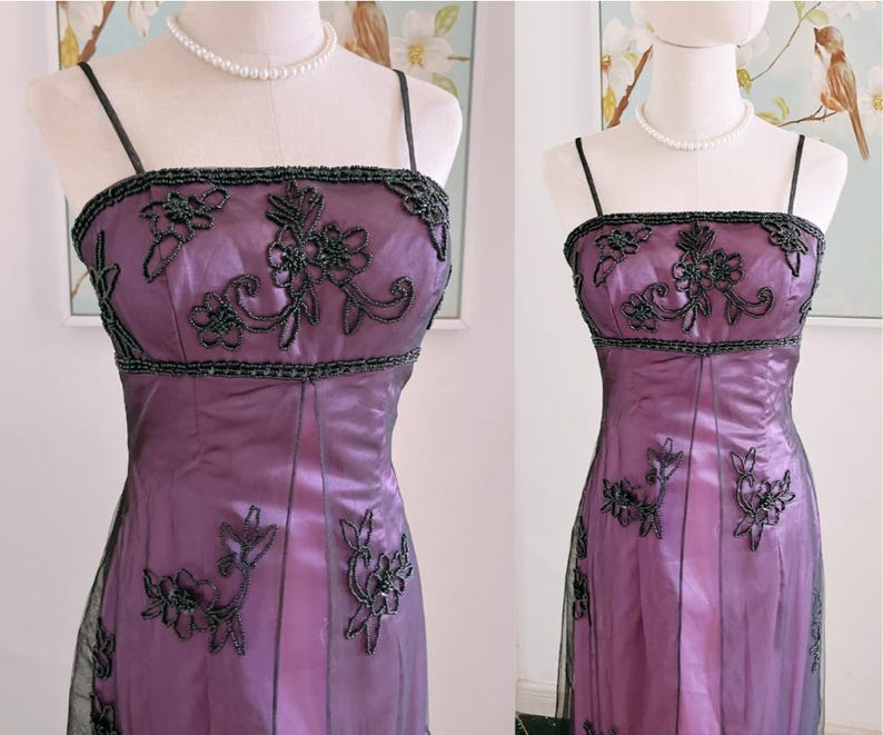 May include: A formal purple dress with black beaded floral embellishments. The dress features a square neckline, spaghetti straps, and a fitted bodice. The skirt is a flowing style, and the dress is displayed on a mannequin.