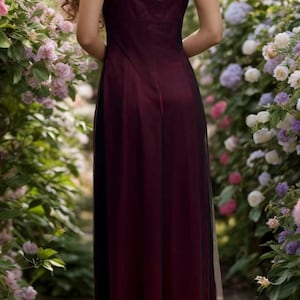 May include: A woman wearing a long, burgundy dress with a black tulle overlay. The dress has a deep V-neckline and a fitted bodice. The woman is standing in a garden with flowers.