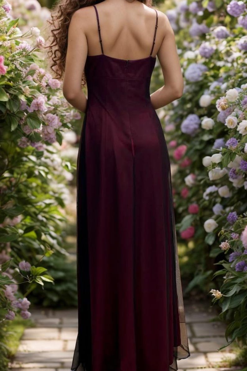 May include: A woman wearing a long, burgundy dress with a black tulle overlay. The dress has a deep V-neckline and a fitted bodice. The woman is standing in a garden with flowers.