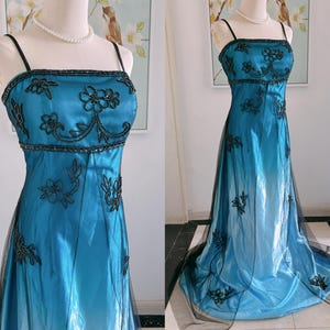 May include: A teal-colored formal gown with black floral embroidery and spaghetti straps. The dress features a fitted bodice with beaded accents and a flowing skirt with a black tulle overlay. The dress is displayed on a mannequin.