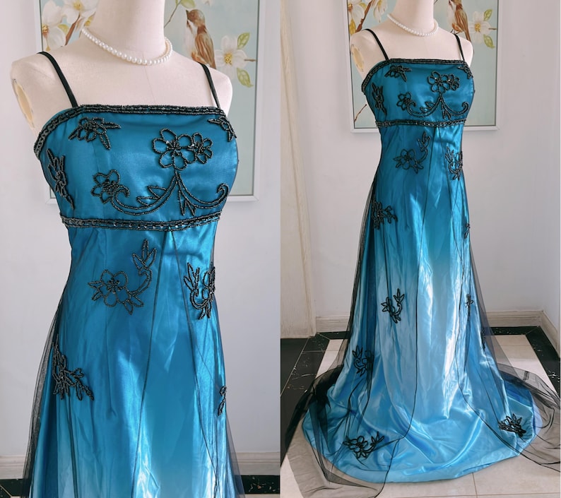 May include: A teal-colored formal gown with black floral embroidery and spaghetti straps. The dress features a fitted bodice with beaded accents and a flowing skirt with a black tulle overlay. The dress is displayed on a mannequin.
