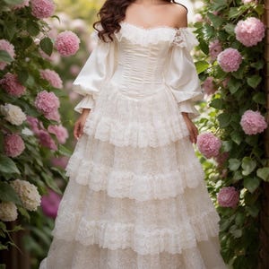 Pode incluir: Um vestido cor de creme com ombros descobertos, apresentando um corpete estilo espartilho, mangas compridas bufantes com acabamento em renda e uma saia longa composta por v&aacute;rias camadas de tecido de renda floral com bainha recortada.