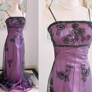 May include: A formal, floor-length purple gown with black beaded embellishments and spaghetti straps. The bodice and skirt feature intricate floral embroidery. A sheer black overlay adds dimension to the skirt.