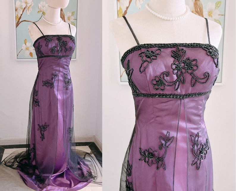 May include: A formal, floor-length purple gown with black beaded embellishments and spaghetti straps. The bodice and skirt feature intricate floral embroidery. A sheer black overlay adds dimension to the skirt.