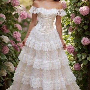 May include: A white off-the-shoulder dress featuring a fitted bodice and a multi-tiered skirt. The garment is constructed with intricate lace detailing, including ruffled lace trim along the neckline and horizontal lace layers on the skirt.