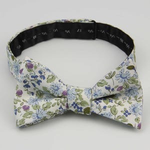 Puede incluir: Una pajarita floral con fondo blanco, con flores azules y verdes y bayas moradas. La banda ajustable para el cuello, de color negro, está marcada con medidas de talla.