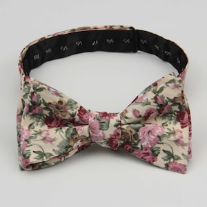 Puede incluir: Una pajarita floral con fondo crema y flores rosas y rojas. La pajarita tiene una correa ajustable negra.