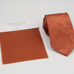 May include: An auburn-colored paisley tie with a matching fabric swatch. The tie is rolled up, displaying its intricate pattern. The fabric swatch is a solid color with a scalloped edge. The text "AZAZIE CHIFFON LINING" and "Auburn" are visible.