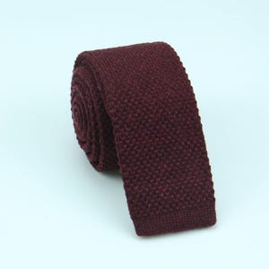Puede incluir: Una corbata de color burdeos intenso con una construcción de punto texturizado. La corbata está enrollada en forma circular en la parte superior, con el extremo plano extendiéndose hacia abajo, mostrando un patrón tejido denso y uniforme.