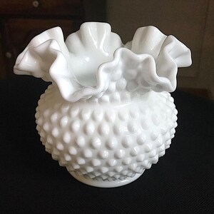 Könnte beinhalten: Eine weiße Hobnail-Vase mit gerüschtem Rand. Die Vase hat einen runden Körper, der mit kleinen, erhabenen Noppen bedeckt ist. Der gerüschte Rand verleiht der Vase im Vintage-Stil eine dekorative Note.