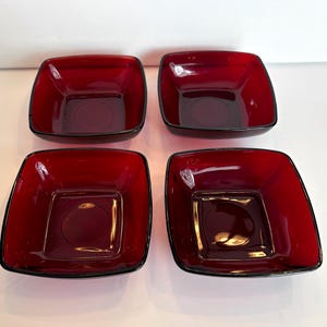 May include: Four square ruby red glass bowls with a black trim. The bowls have a slightly rounded square shape with a circular indentation in the center. The bowls are arranged in a square formation.