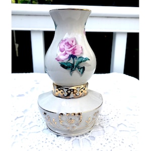 Puede incluir: Un jarrón de cerámica blanco con un borde dorado y un diseño de rosa rosa. El jarrón tiene forma de lámpara con una base redonda.