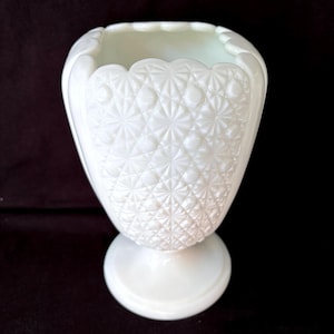 Pode incluir: Um vaso branco, de estilo vintage, com um padrão texturizado de diamantes e estrelas. O vaso tem uma borda ondulada e uma base redonda. O vidro opaco tem uma aparência leitosa. O vaso é provavelmente usado para fins decorativos.