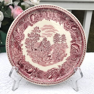 May include: A vintage, white ceramic saucer with a red floral pattern. The design features a cottage in a wooded landscape with a boat on a lake. The saucer has a scalloped edge.