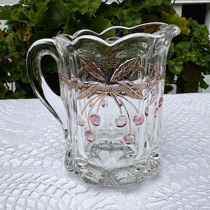 Puede incluir: Jarra de cristal transparente con asa, con un diseño decorativo de cerezas rosas y hojas doradas. La jarra tiene un borde festoneado y una banda dorada. El diseño se repite alrededor del cuerpo.