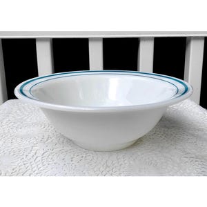 May include: A white bowl with a rounded shape and a teal blue double stripe around the rim. The bowl is sitting on a white, lacy surface. The background is a white wooden structure.