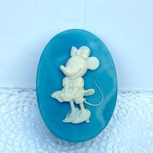May include: An oval-shaped blue soap bar featuring a raised, cream-colored relief of a cartoon mouse character wearing a bow, a dress, and shoes. The character is depicted in a standing pose with a long tail curving to the right side of the oval.