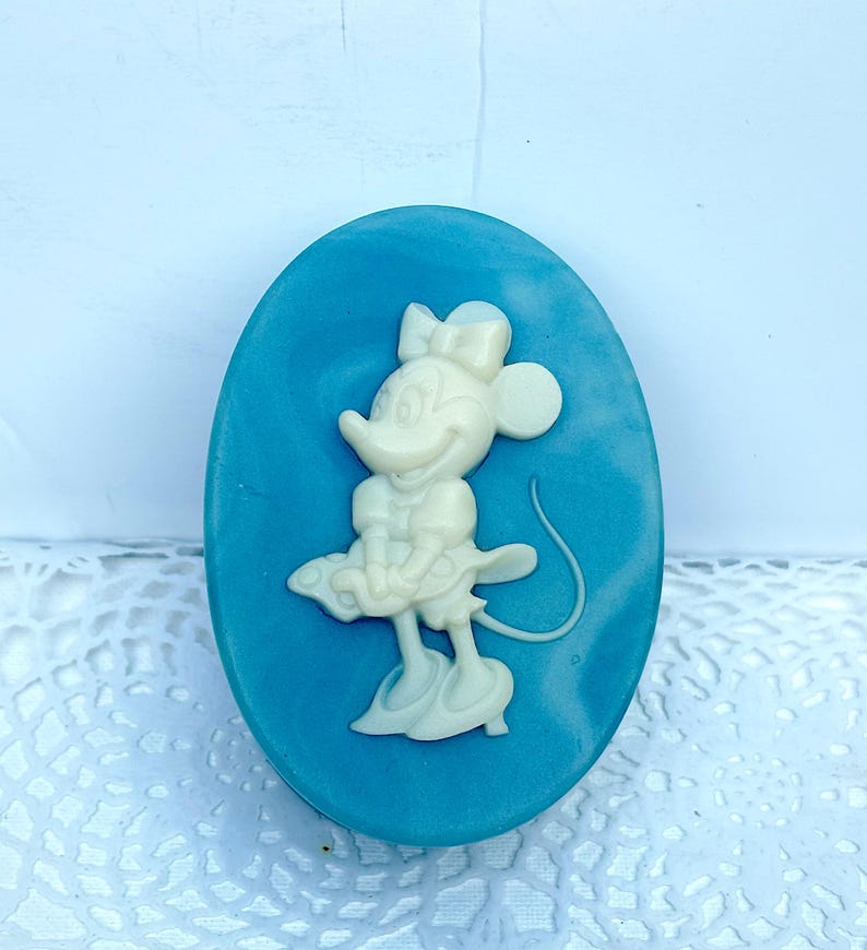 May include: An oval-shaped blue soap bar featuring a raised, cream-colored relief of a cartoon mouse character wearing a bow, a dress, and shoes. The character is depicted in a standing pose with a long tail curving to the right side of the oval.