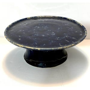 May include: A circular ceramic cake stand featuring a deep navy blue glaze with a mottled white and light blue marbled pattern across the flat top surface. The stand has a wide, flared pedestal base with a smooth, dark navy blue finish.