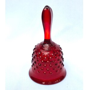 Puede incluir: Una campana de cristal rojo rubí translúcido que presenta una textura de puntos en relieve por todo el exterior del cuerpo. El objeto tiene un mango liso y alargado que se extiende desde la parte superior de la base redondeada y acampanada.