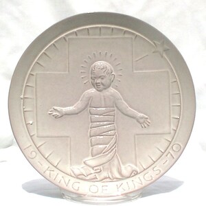 Könnte beinhalten: Ein weißer Keramikteller mit einem Reliefbild eines Jesuskindes in einem weißen Gewand, das auf einem Kreuz steht. Der Teller hat einen weißen Rand mit den Worten "King of Kings" um den Rand herum.