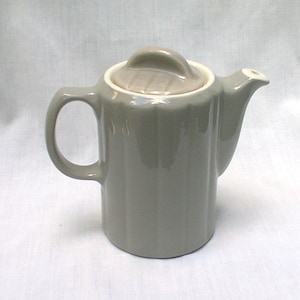 May include: A gray ceramic teapot with a matching lid. The teapot has a ribbed design and a curved handle. The lid has a rounded handle. The spout is small and curved. The teapot is on a white surface.